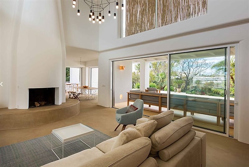 Salón de doble altura con chimenea de obra y ventanales al jardín en Aloha
