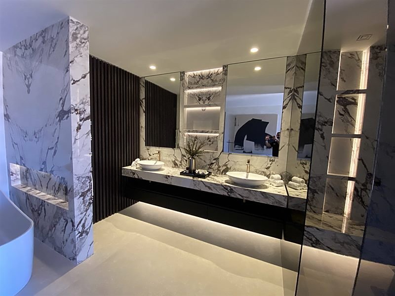 Baño principal revestido en mármol veteado, con doble lavabo e iluminación indirecta